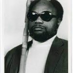 John Langston Gwaltney headshot in a black suit and white turtle neck with his cane showing.