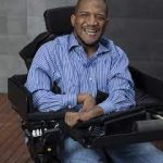 Lateef McLoud wearing jeans and a blue shirt in his wheelchair smiling.