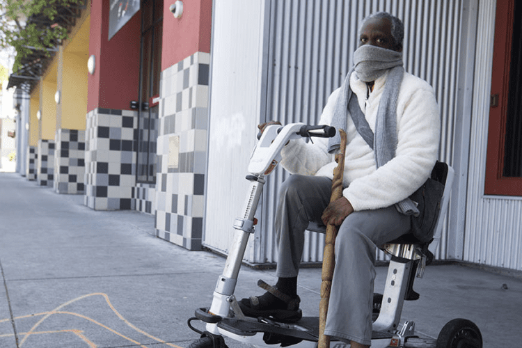 Leroy sitting in his scooter wearing a white sweater and grey pants.